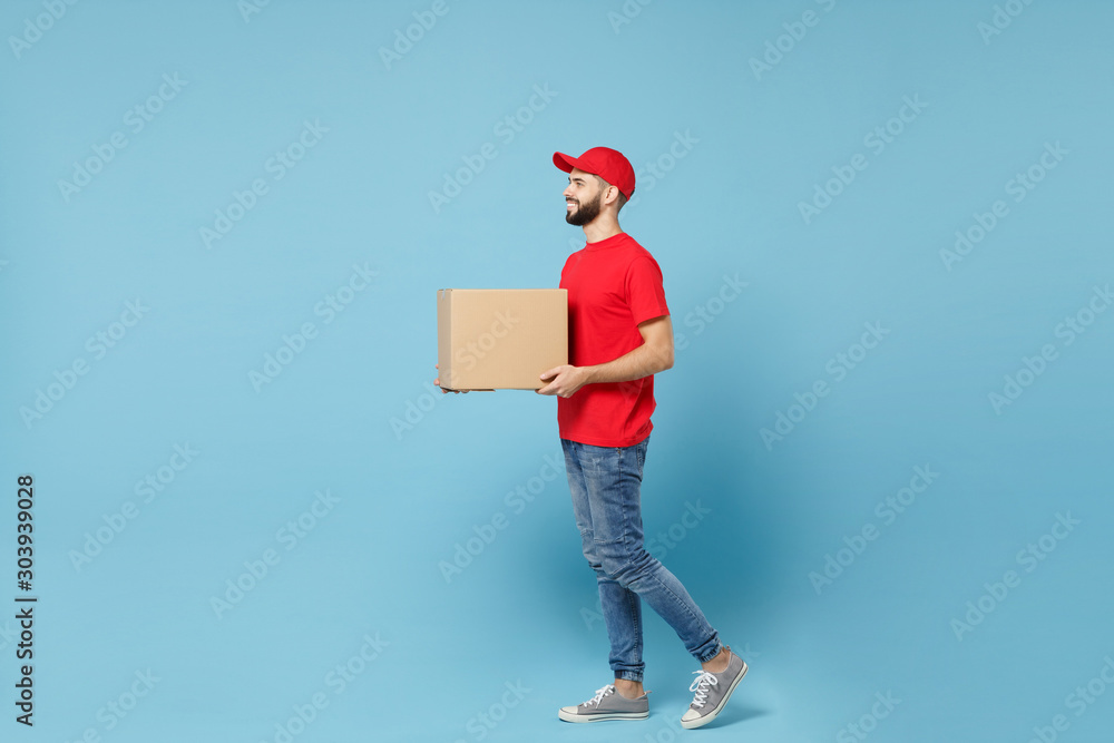 Delivery man in red uniform isolated on blue background, studio portrait. Male employee in cap t-shirt print working as courier dealer hold empty cardboard box. Service concept. Mock up copy space. Delivery man in red uniform isolated on blue background, studio portrait. Male employee in cap t-shirt print working as courier dealer hold empty cardboard box. Service concept. Mock up copy space.