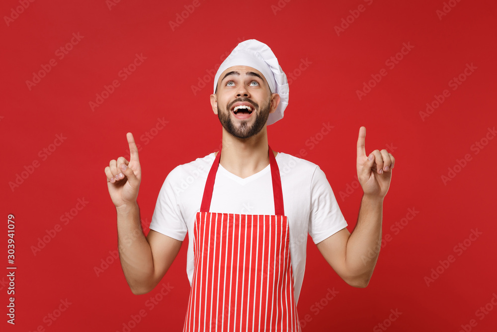 Amazed young bearded male chef cook baker man in striped apron white t-shirt toque chefs hat posing isolated on red background. Cooking food concept. Mock up copy space. Pointing index fingers up. Amazed young bearded male chef cook baker man in striped apron white t-shirt toque chefs hat posing isolated on red background. Cooking food concept. Mock up copy space. Pointing index fingers up.