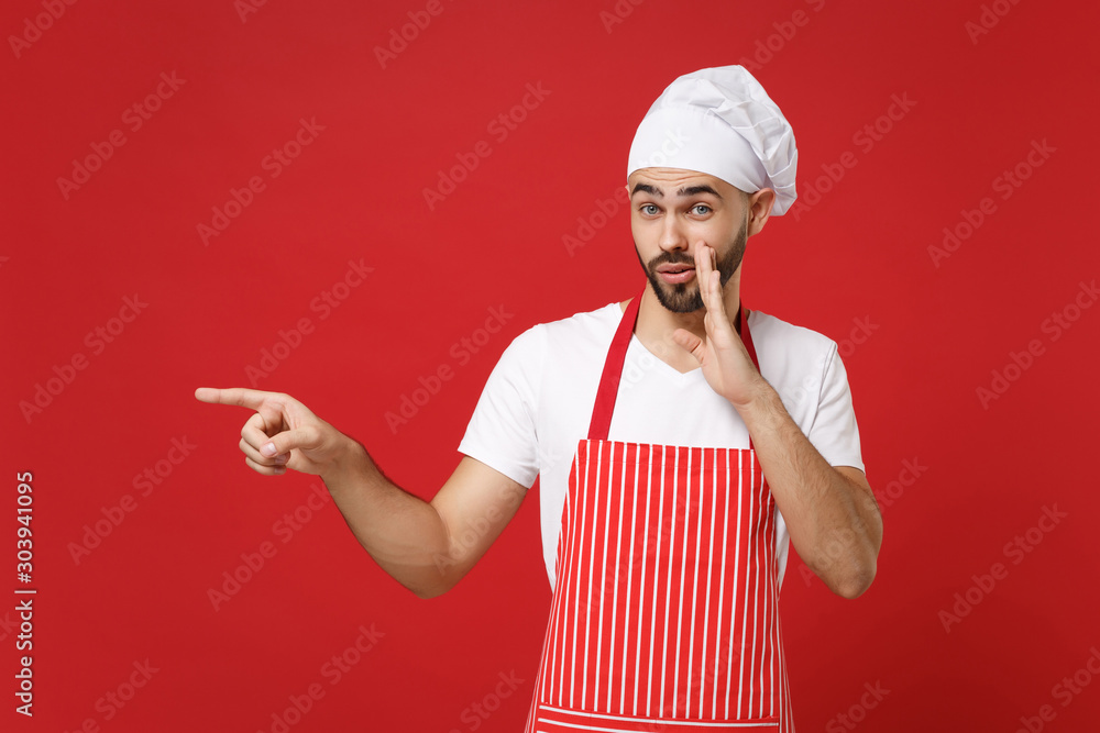 Secret chef cook baker man in apron toque chefs hat posing isolated on red background. Cooking food concept. Mock up copy space. Whispers gossip, tells secret with hand gesture, pointing finger aside. Secret chef cook baker man in apron toque chefs hat posing isolated on red background. Cooking food concept. Mock up copy space. Whispers gossip, tells secret with hand gesture, pointing finger aside.