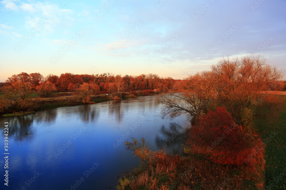 Brązowe i czerwone drzewa nad rzeką w świetle zachodzącego słońca. - obrazy, fototapety, plakaty 