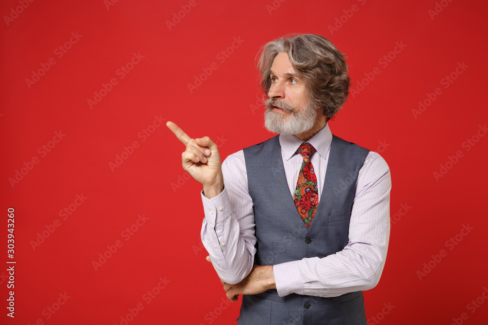 Pensive elderly gray-haired mustache bearded man in classic shirt vest and colorful tie isolated on red background in studio. People lifestyle concept. Mock up copy space. Pointing index finger aside. Pensive elderly gray-haired mustache bearded man in classic shirt vest and colorful tie isolated on red background in studio. People lifestyle concept. Mock up copy space. Pointing index finger aside.