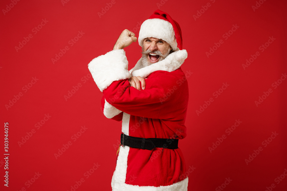 Obraz premium Side view of elderly gray-haired mustache bearded Santa man in Christmas hat posing isolated on red background. New Year 2020 celebration holiday concept. Mock up copy space. Showing biceps, muscles.