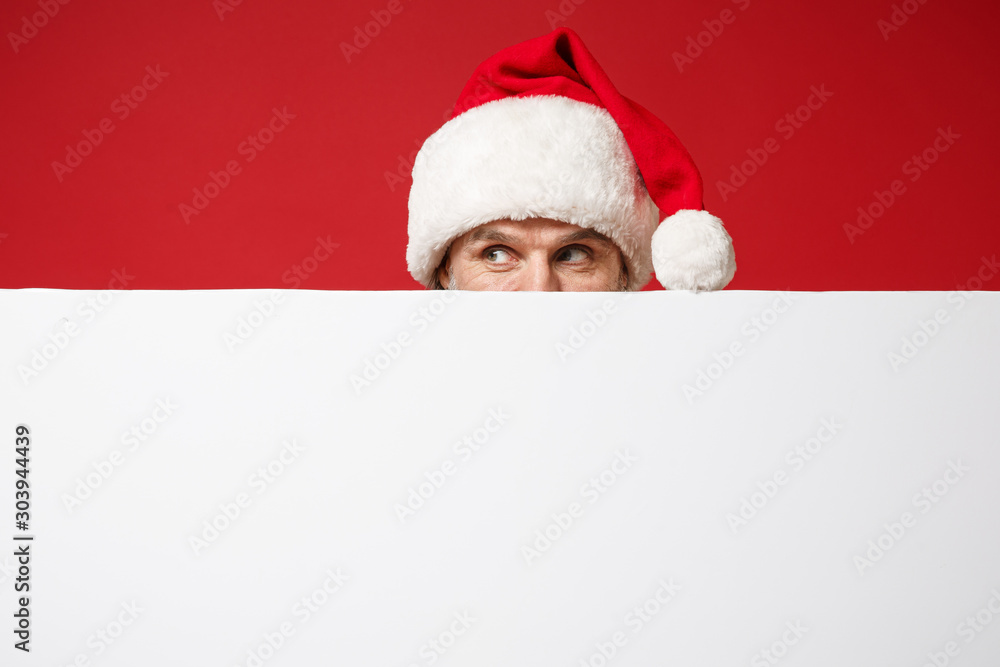 Elderly gray-haired mustache bearded Santa man in Christmas hat posing isolated on red background. New Year 2020 celebration holiday concept. Mock up copy space. Covering with white blank billboard. Elderly gray-haired mustache bearded Santa man in Christmas hat posing isolated on red background. New Year 2020 celebration holiday concept. Mock up copy space. Covering with white blank billboard.