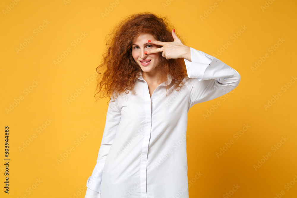 Smiling young redhead woman girl in casual white shirt posing isolated on yellow orange wall background in studio. People sincere emotions lifestyle concept. Mock up copy space. Showing victory sign. Smiling young redhead woman girl in casual white shirt posing isolated on yellow orange wall background in studio. People sincere emotions lifestyle concept. Mock up copy space. Showing victory sign.