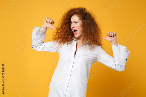 Cheerful young redhead woman girl in casual white shirt posing isolated on yellow orange wall background  studio portrait. People lifestyle concept. Mock up copy space. Pointing thumbs on herself.