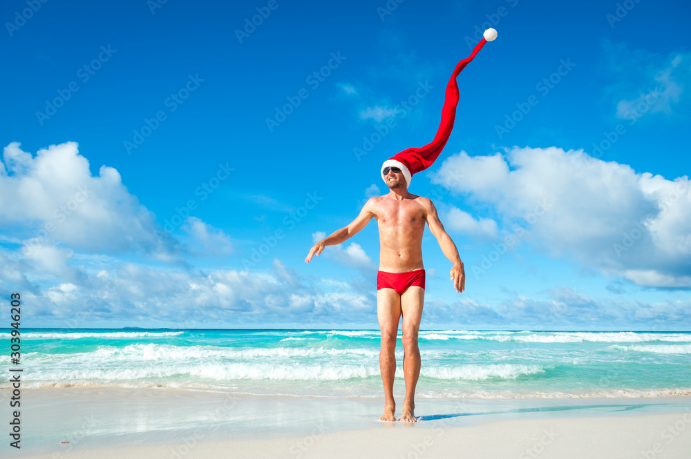 Fit man with extra long Santa hat celebrating the holidays in Christmas red swimming briefs on the shore of a tropical beach Fit man with extra long Santa hat celebrating the holidays in Christmas red swimming briefs on the shore of a tropical beach