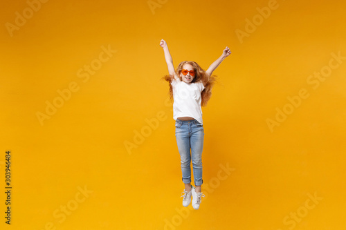 Cheerful little ginger kid girl 12-13 years old in white t-shirt, heart glasses isolated on yellow wall background. Childhood lifestyle concept. Mock up copy space. Having fun, jumping, rising hands.