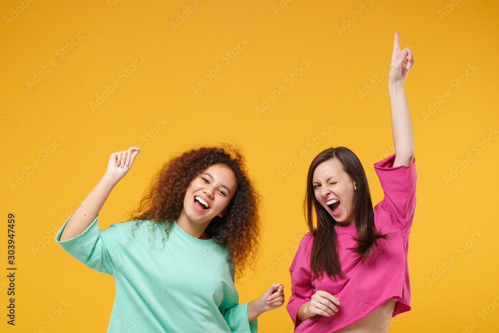 Two cheerful women friends european african american girls in pink green clothes posing isolated on yellow background. People lifestyle concept. Mock up copy space. Having fun dancing rising hands up. - obrazy, fototapety, plakaty 