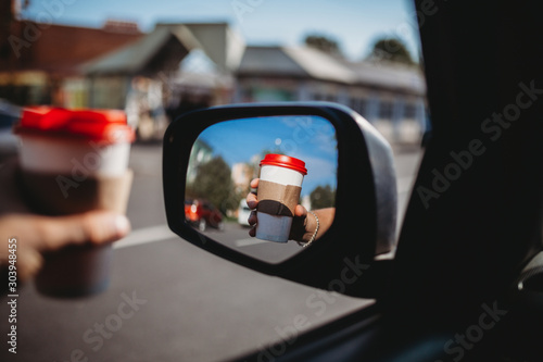 Close-up of the driver leaning out the window hand holding a paper white coffee Cup with a red lid. Hand with a Cup of hot tea in the reflection of the rear view glass. Toned image