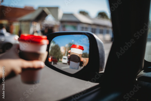 Close-up of the driver leaning out the window hand holding a paper white coffee Cup with a red lid. Hand with a Cup of hot tea in the reflection of the rear view glass. Toned image
