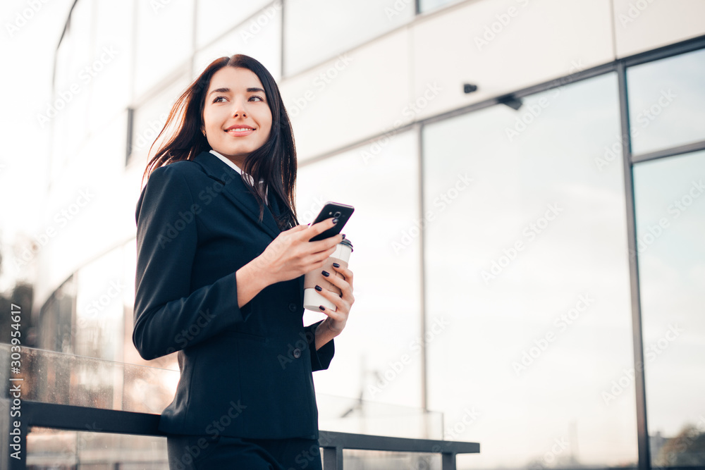 Successful smiling businesswoman or entrepreneur using phone and drink coffee standing in front of his office.