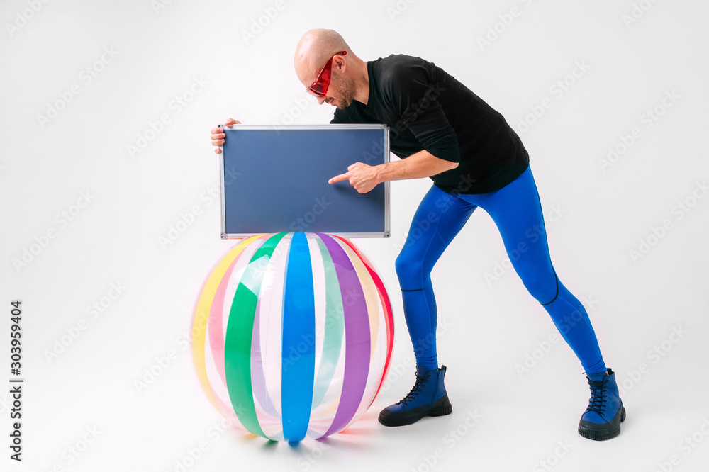 Athlete bald man in red sunglasses and blue sportive tights standing near beach ball and showing his hand to nameplate. Sport motivation concept.