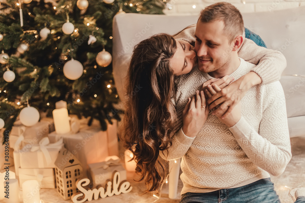 Fototapeta premium Beautiful young couple kisses sitting on the floor and celebrating new year in front of Christmas tree.