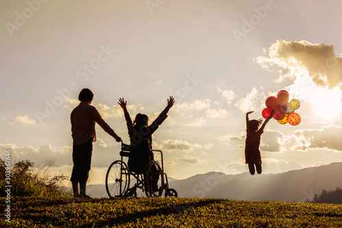 Little girl jumping holding multicolored balloons, mother in wheelchair with raised arms looking at sunset. happy family concepts.