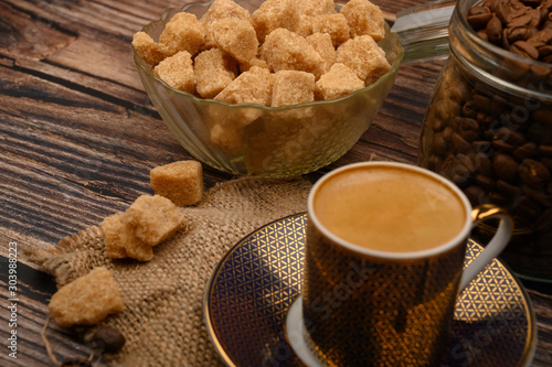 A Cup of coffee, pieces of brown sugar in a sugar bowl, coffee beans in a glass jar on a wooden background.