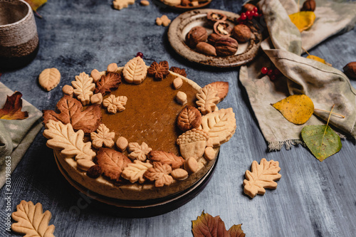 Fototapeta Naklejka Na Ścianę i Meble -  traditional pumpkin pie decorated with leaves shaped cookies on shabby blue background with coffee and textile