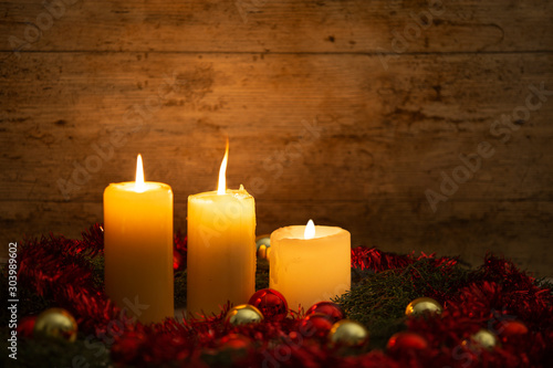 The warmth of the Christmas concept: three candles lit on a light wooden table and a rustic setting with pine branches, red decoration and gold and red bright baubles with bokeh effect