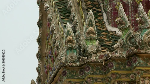 A daylight closeup shot of intricate chofa guardians statue made of porcelain placed against the vivid foliage ornamentation of the roof of Phra Sawet Kudakhan Wihan Yot in Thailand. photo