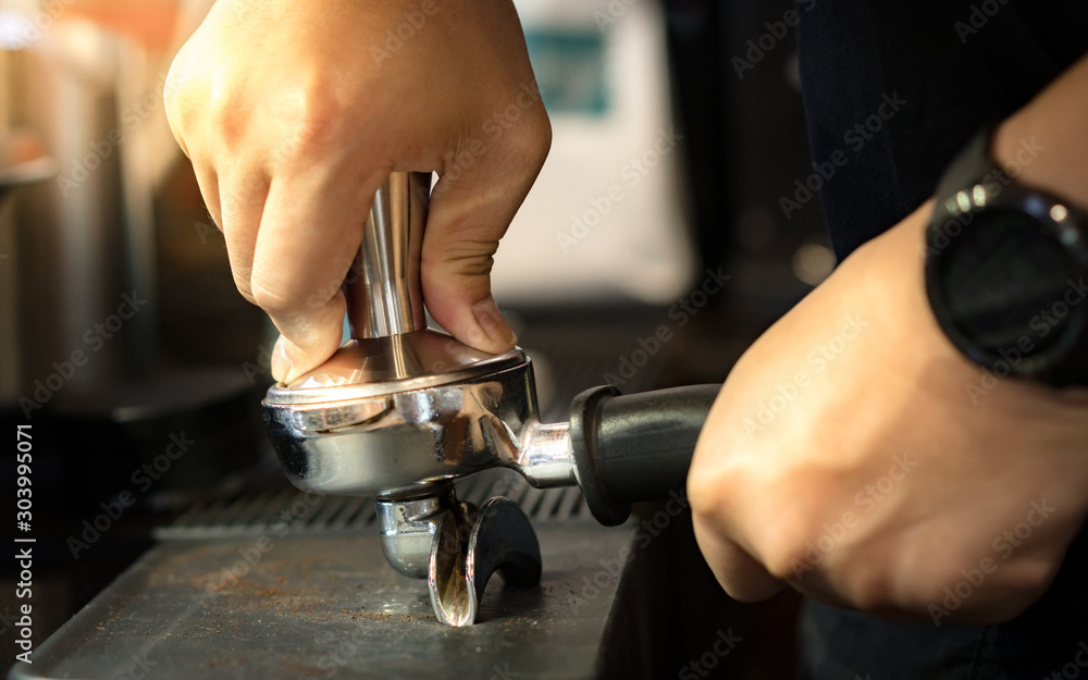 Closeup, barista's right hand hold coffee tamper and tamp arabica grounds  in espresso double spout portafilter gripped by left on tamp mat before  brewing with espresso machine. Practice makes perfect. Stock-Foto