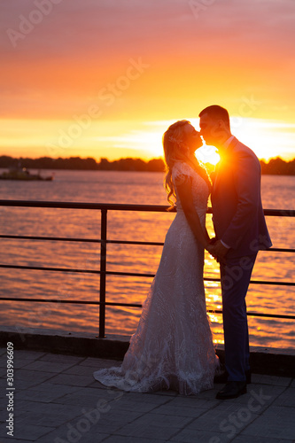 Young beautiful couple of newlyweds with bride and groom walk on the terrace by the ocean during a wedding ceremony in a wedding dress and suit kissing and hugging at sunset. Love and lovestory.