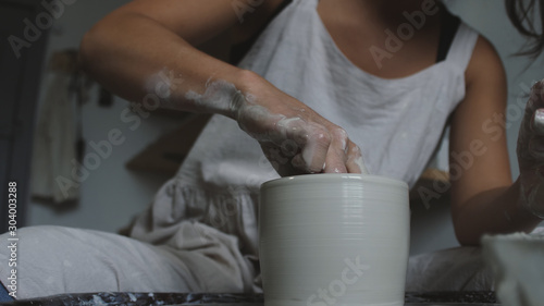 Master female potter molding wet clay into a bowl with wet sponge on potter's wheel. Female potter works in studio.