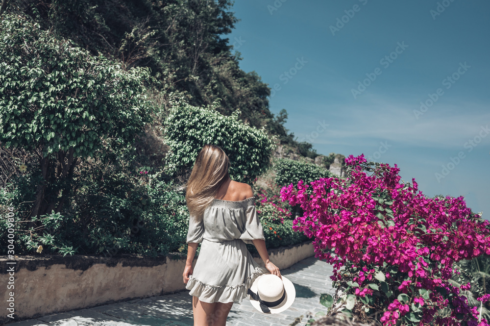 Fototapeta premium Beautiful blonde woman back view walks and admires tropical landscapes at paradise island hotel in Sanya, China. Luxury vacation banner. Tropical resort concept. Asia travel lifestyle tourist