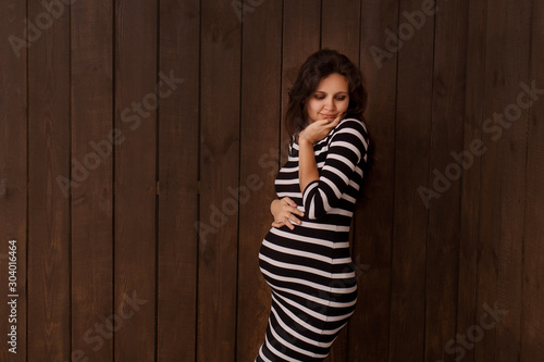 Pregnant woman in stripe dress holds hands on belly on a dark brown background. Pregnancy, maternity, preparation and expectation concept. Beautiful tender mood photo of pregnancy © vik_li