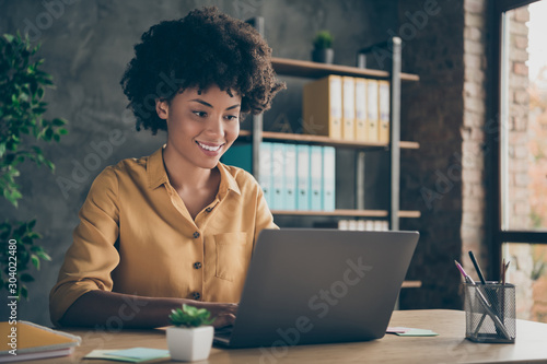 Photo of cheerful positive mixed-race girl smiling toothily working on presentation about her corporation using laptop on desktop