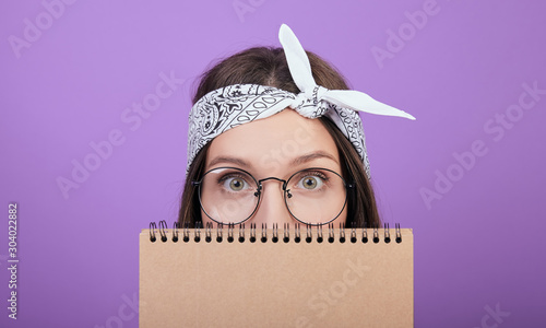 Fotografía A cute brown-haired woman in round glasses hides her face behind a notebook