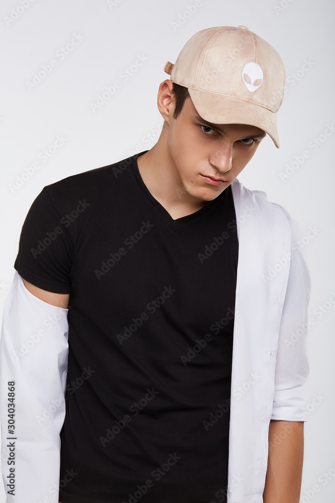 Cropped front view shot of a dark-haired man, wearing beige velvet baseball cap with alien print, black t-shirt and white shirt. He is posing on grey background and looking straight. Cropped front view shot of a dark-haired man, wearing beige velvet baseball cap with alien print, black t-shirt and white shirt. He is posing on grey background and looking straight.