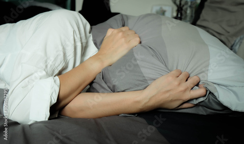 Asian woman wearing white pajamas on black bed use 2 hands to hold press the pillow to cover face and head. To prevent the light from the window in the morning. Don't want to wake up or all disturb