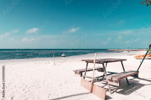An old wooden table type outdoor bench placed on the white sand beach. Background is deep blue and soft sea with 2-3 small boats parked. The clear sky not much clouds. Taken at Maldives  vintage tone.