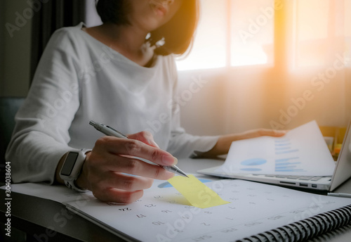 Asian woman is writing on yellow paper notes and put a warning on calendar. The other hand hold the report graph and has laptop on the front. Maybe preparing data for marketing report. window light