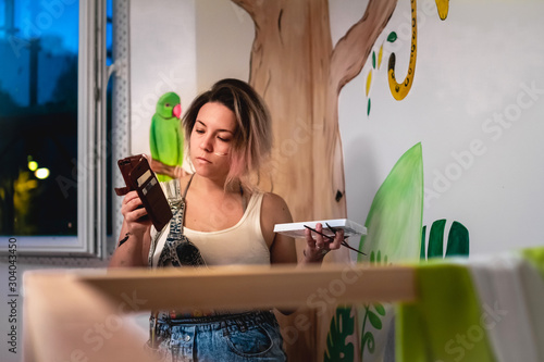 Young female artist searching an inspiration on the social media while painting the wall in the children's room