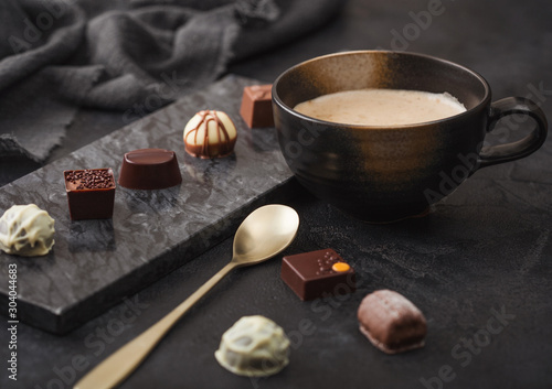 Luxury Chocolate candies selection with cup of cappuccino coffee and dessert spoon on black marble board and dark table background.