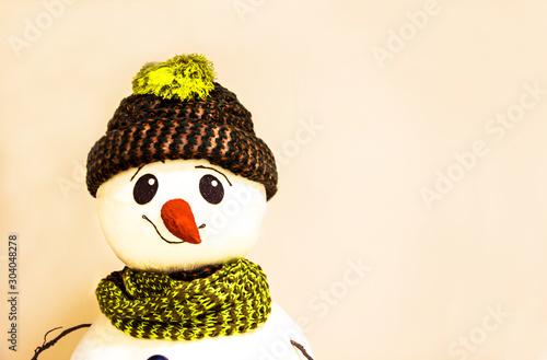 Portrait of a funny - cheerful snowman dressed in a gray-green hat with a scarf on his neck on a beige-pink background.