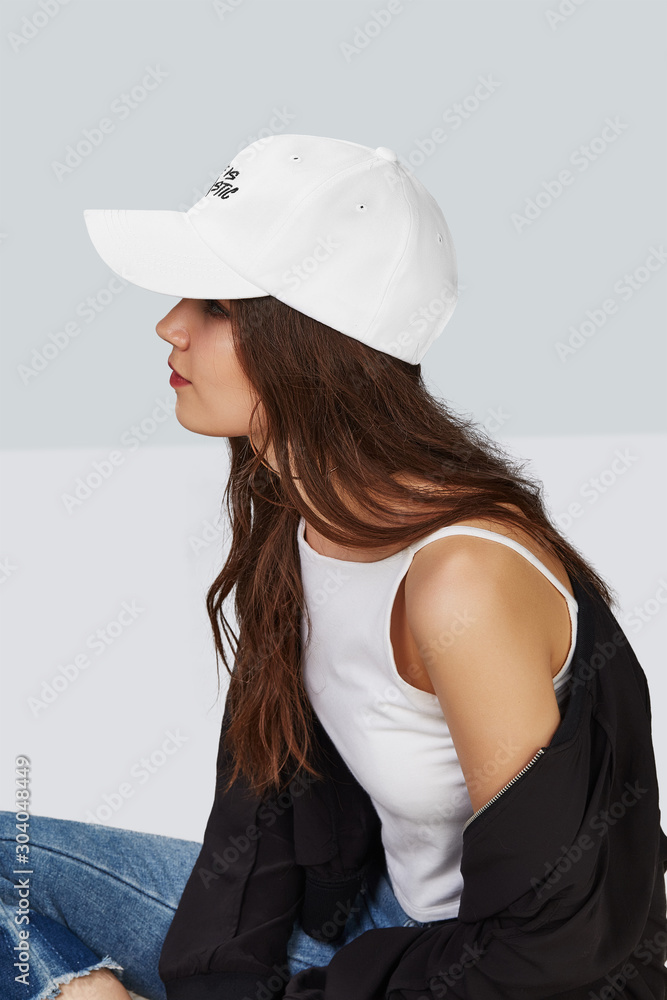 Cropped side photo of a girl, sitting on a floor and wearing white baseball cap with print with lettering Cropped side photo of a girl, sitting on a floor and wearing white baseball cap with print with lettering
