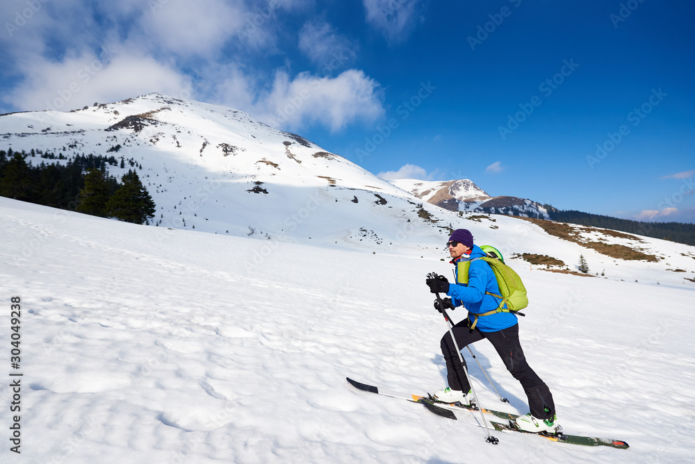 Male tourist in colorful clothing and sunglasses with backpack trekking on skis in deep snow on background of bright blue sky and beautiful mountain. Winter vacations, ski touring concept.