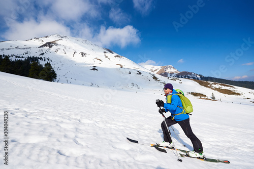 Male tourist in colorful clothing and sunglasses with backpack trekking on skis in deep snow on background of bright blue sky and beautiful mountain. Winter vacations, ski touring concept.