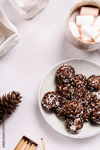 Swedish chocolate balls and cup of cocoa with marshmallow on grey concrete table. Cozy winter breakfast. Chokladbollar. Homemade healthy raw cocoa oatmeal balls sprinkled with coconut flakes