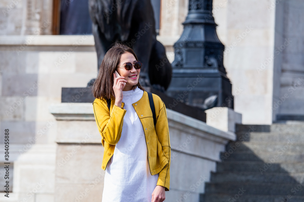 Fototapeta premium Portrait of a young, attractive and beautiful Asian woman smiling as she talks on her smartphone int he city. She is looking away as she talks animatedly and happily on her mobile device.