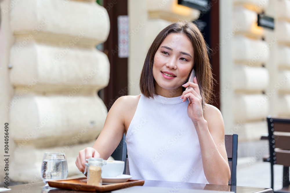 Smiling Asian woman drinking coffee and using her mobile phone. Satisfied female enjoying cup of coffee. Close up portrait of beautiful girl drinking coffee from a white mug in the coffee shop Smiling Asian woman drinking coffee and using her mobile phone. Satisfied female enjoying cup of coffee. Close up portrait of beautiful girl drinking coffee from a white mug in the coffee shop