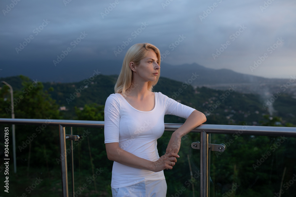 Blonde caucasian woman in standing near the edge of observation deck viewpoint or balcony with metallic handrails and transparent glass walls looking towards mountains in evening view from back Blonde caucasian woman in standing near the edge of observation deck viewpoint or balcony with metallic handrails and transparent glass walls looking towards mountains in evening view from back