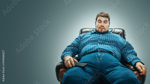 Portrait of fat caucasian man wearing jeanse and whirt sitting in a brown armchair isolated on gradient grey background. Emotional watching TV and changing channels, laughting. Overweight, carefree.