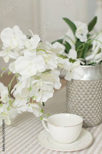 A white empty cup with plate on white tablecloth on white bright background with white flowers of orchid and green leaves like wedding morning