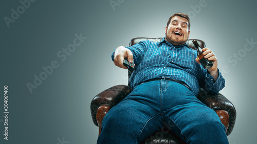 Portrait of fat caucasian man wearing jeanse and whirt sitting in a brown armchair isolated on gradient grey background. Watching TV drinks beer and changing channels, laughting. Overweight, carefree.