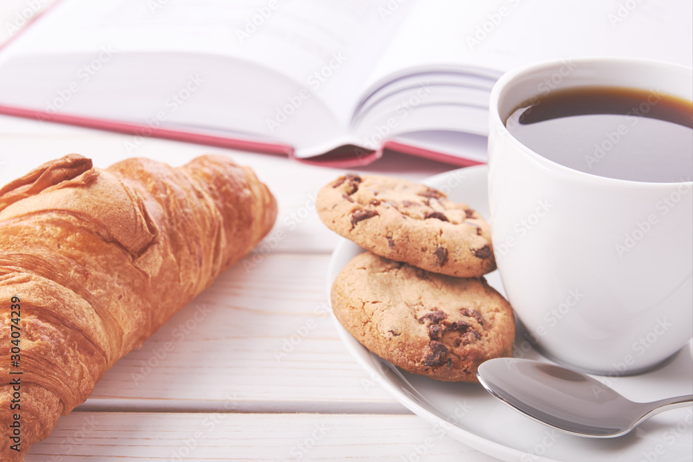 Still life with red book, cup of coffee or tea and two cookies. Croissant on white table top. Still life with red book, cup of coffee or tea and two cookies. Croissant on white table top.