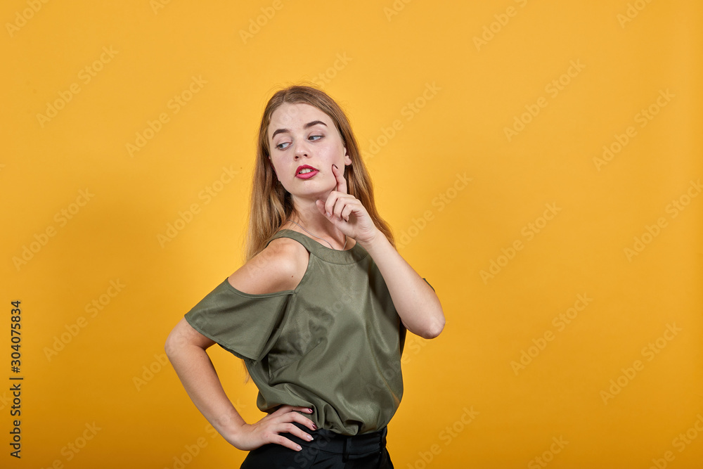 Portrait of shocked young woman in casual clothes keeping mouth wide open, keeping finger on cheek isolated on orange wall background in studio. People sincere emotions lifestyle concept. Portrait of shocked young woman in casual clothes keeping mouth wide open, keeping finger on cheek isolated on orange wall background in studio. People sincere emotions lifestyle concept.