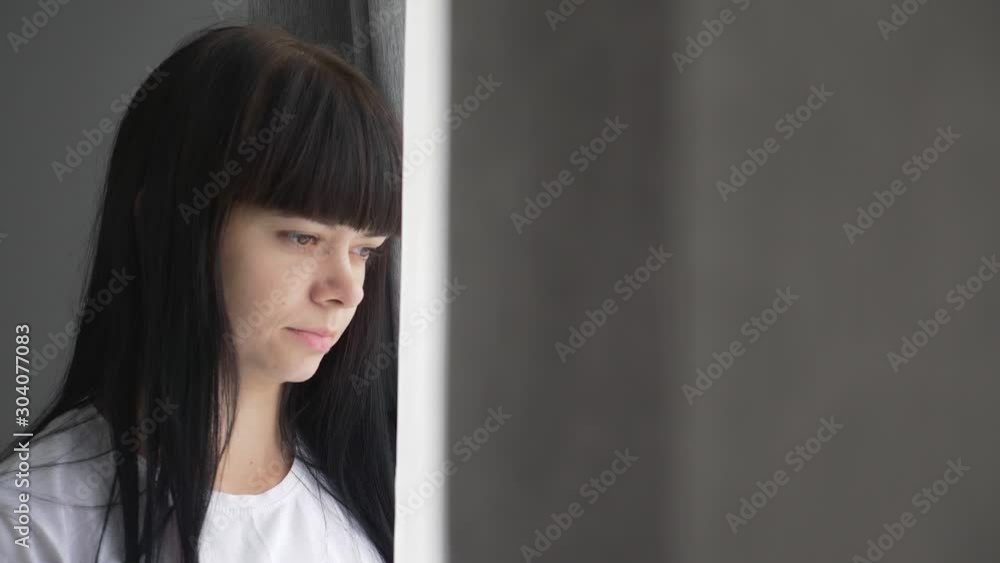 Attractive dark-haired young woman looks out the window at the cityscape. She walks through a large bright room and goes to the curtains. Attractive dark-haired young woman looks out the window at the cityscape. She walks through a large bright room and goes to the curtains.