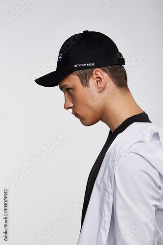 Cropped side photo of a dark-haired man, wearing black baseball cap with Chinese characters print and lettering "be your muse" on the left side of a cap, black t-shirt and white shirt.  © RedUmbrella&Donkey